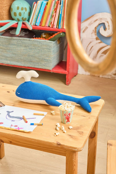 Blue plush whale toy on a wooden table with popcorn and colouring books in a child's room.