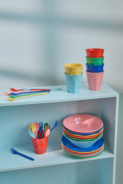 Colourful children's tableware including cups, plates, and utensils on a shelf against a light blue wall.