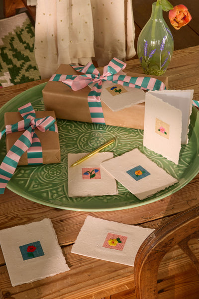 Gifts wrapped in brown paper with colourful ribbons on a green plate, surrounded by handmade cards with small  floral woven designs
