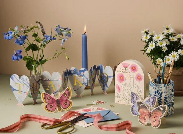 A styled tabletop arrangement with decorative paper butterflies, floral heart-shaped cards spelling 'THANK YOU', a blue candle, flowers, scissors, ribbon, and stationery holders against a warm beige background.