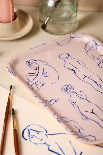 Pink tray with blue line drawings of women, surrounded by paintbrushes and a glass of water on a beige surface.