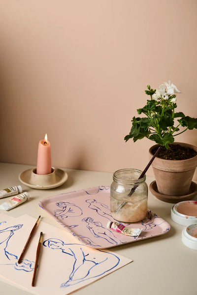 Artistic setup with a tray, candle, plant and art materials on a table against a beige wall.