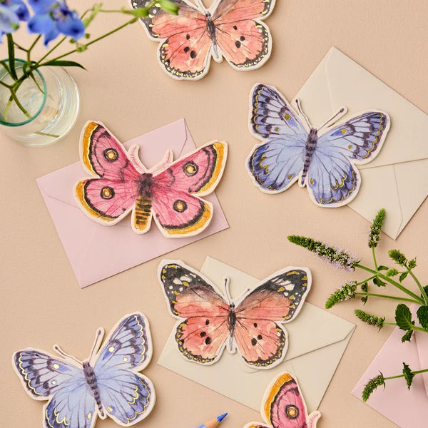 A styled flat-lay of butterfly-shaped cards arranged on pastel envelopes and floral props against a warm beige background. The butterflies feature watercolor-style designs in pink, peach, lavender, blue, and black details.