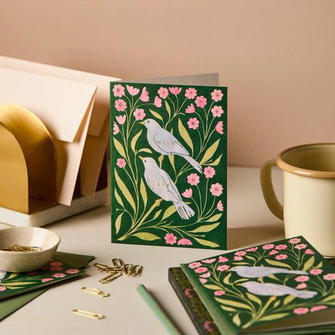 Decorative greeting card with birds and flowers on a desk with stationery items.