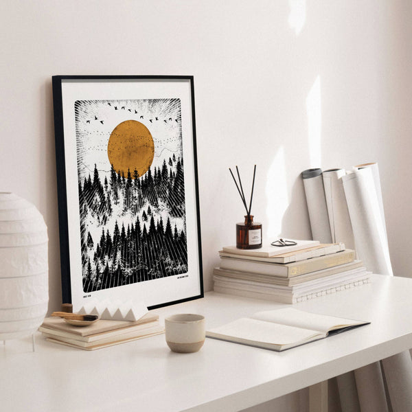 Framed artwork of a forest and sun on a white desk with books and decor.