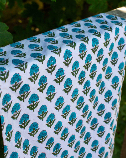 A close-up of a white tea towel, featuring a repeating block-print style pattern of teal flowers and green leaves, draped outdoors against a blurred green foliage background.