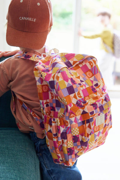 A child wearing a colourful checkered backpack and an orange cap indoors.