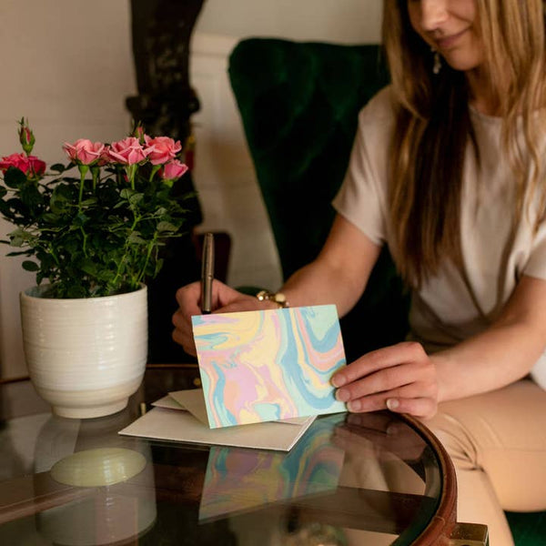 Woman writing in a colourful marbled card next to a potted plant on a reflective surface.