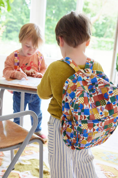 Child with a colourful geometric-patterned backpack standing at a table with another child.