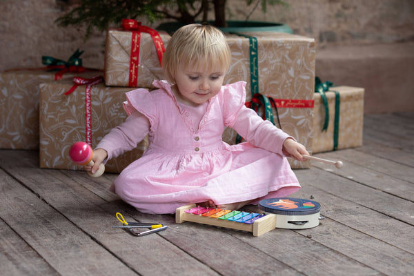 Child in a pink dress playing with musical instruments on a wooden floor with wrapped gifts in the background.