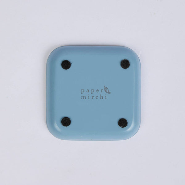 The back of a light blue square enamelled iron coaster with rounded corners and four black circular feet, featuring the 'paper mirchi' logo printed in the centre, on a clean white background.