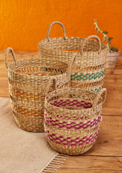 Three woven natural-fibre storage baskets with handles, in large, medium and small, decorated with horizontal bands of green, yellow, and magenta. They are styled on a beige rug over a wooden floor against a textured orange wall, with a small potted plant in the background.