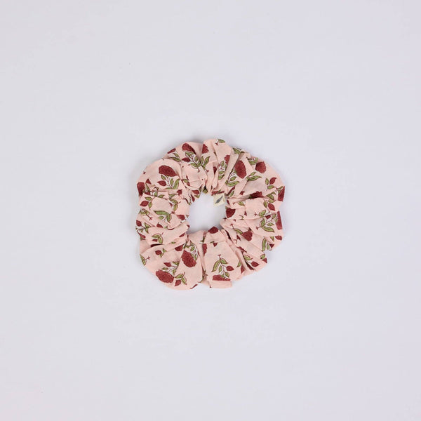 A fabric hair scrunchie with a light pink background and a red flower and green leaf pattern, centred on a plain white background.