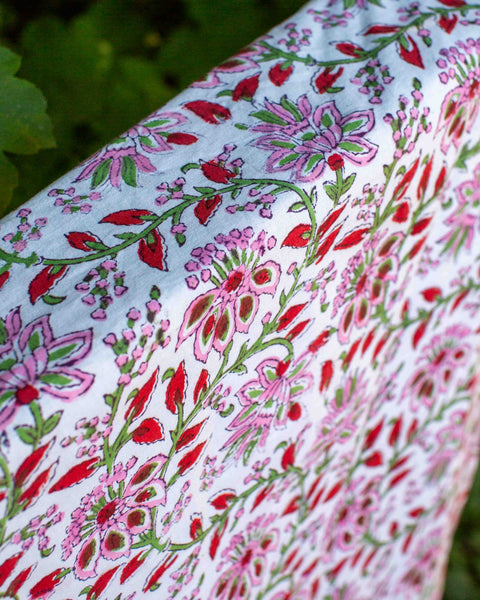 A close-up of a tea towel with a colourful floral block print pattern in pink, red, and green, draped outdoors with blurred green foliage in the background.