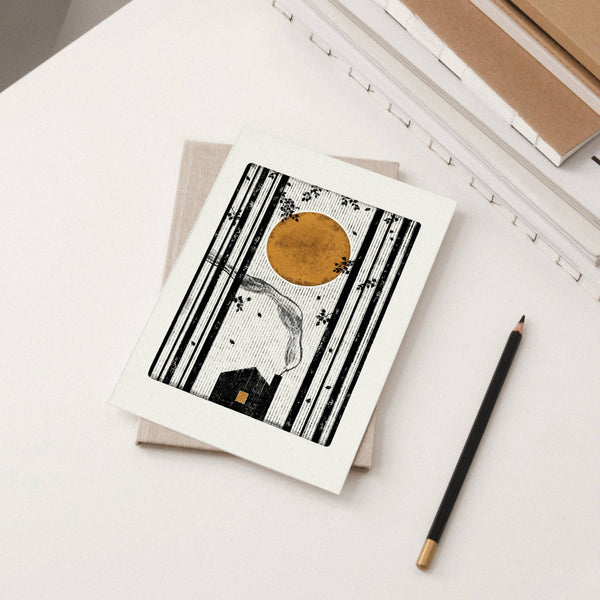 Greeting card with black and white design featuring a moon and house, next to a pencil on a light surface.
