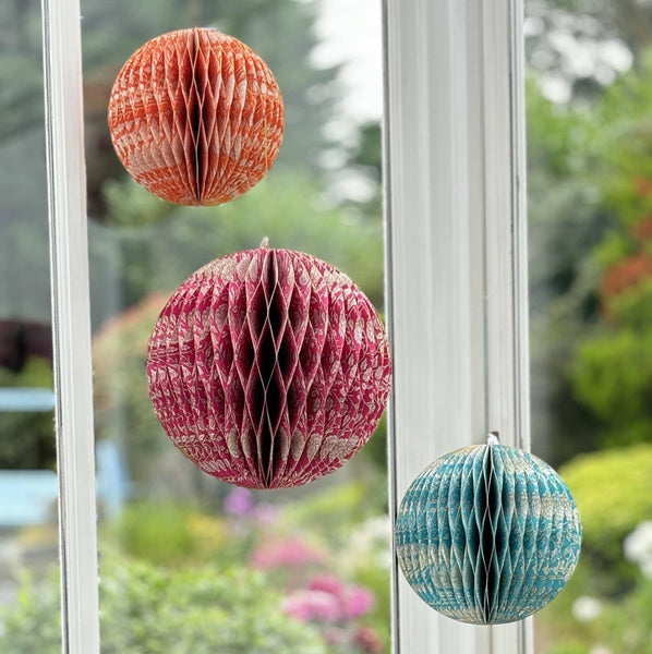 Three hanging honeycomb paper lanterns in orange, pink, and blue are displayed on a white window with a blurred garden background.