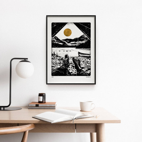 Framed artwork of a people sitting in a tent by a lake with mountains, placed above a desk.