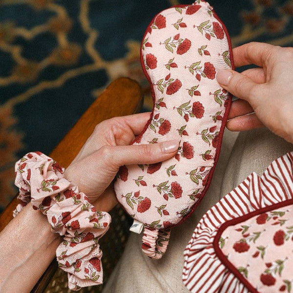 A close-up of a person holding a light pink fabric eye mask with a red flower and green leaf pattern, with matching scrunchie and striped pillowcase also visible.