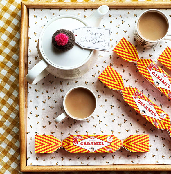 A styled flat-lay of Christmas tea treats on a wooden tray with a patterned cloth. Visible items include a white teapot with a chocolate mug on top, two cups of tea, and several orange-and-yellow striped Tunnock's Christmas crackers.