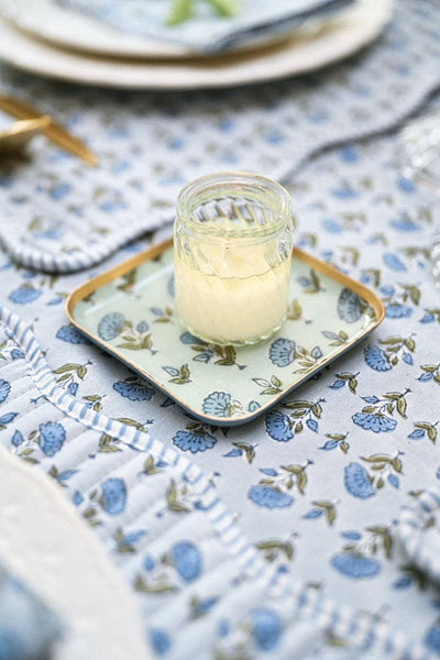 A small clear glass jar filled with pale yellow cream sits on a square decorative coaster/tray with a blue and green floral pattern, placed on a matching blue floral tablecloth in a styled tabletop setting.
