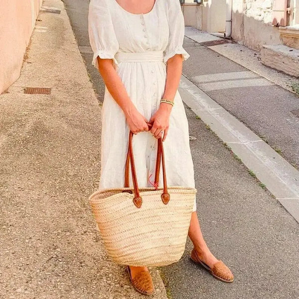 Woman in a white dress holding a straw bag on a street.