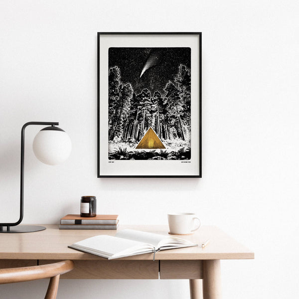 Framed artwork of a tent in a forest with a shooting star above, displayed on a wall above a desk.