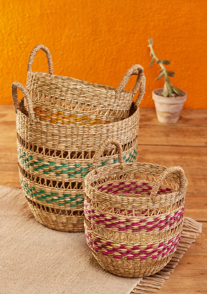 Three woven natural-fibre storage baskets with handles, in large, medium and small, decorated with horizontal bands of green, yellow, and magenta. They are styled on a beige rug over a wooden floor against a textured orange wall, with a small potted plant in the background.