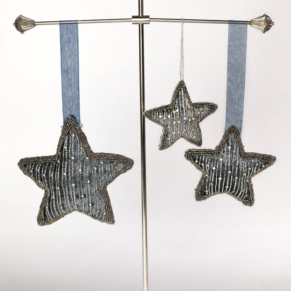Three star-shaped beaded ornaments in a distressed blue-grey colour, hanging from a silver metal display stand against a white background.