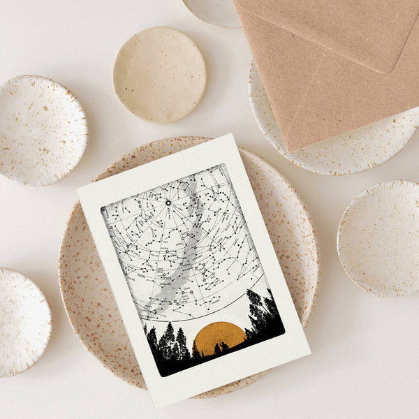 Greeting card featuring a star map with trees and a sunset on a white surface with ceramic plates.