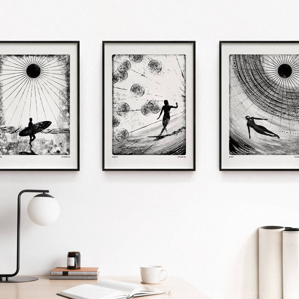Three framed black and white artworks of surfers on a wall with a lamp and books on a desk below.