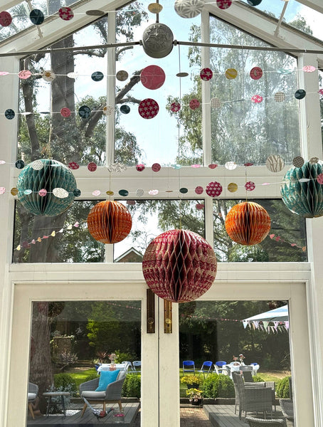 Colourful hanging honeycomb paper lanterns and circular string banners decorated in a sunroom with large windows overlooking a garden patio.