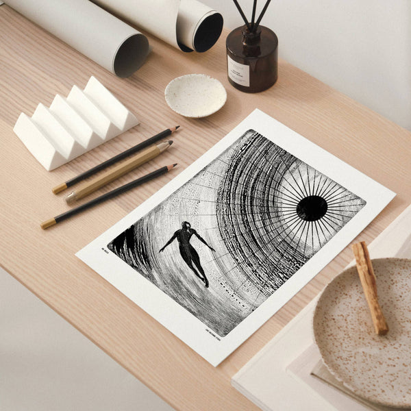 Art print of a black and white silhouette of a person swimming in water under a radial sun on a wooden table with art supplies.