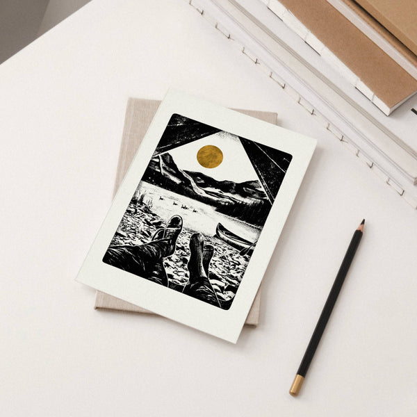 Artistic greeting card of a people sitting by a lake with a golden sun, placed on a light surface with a pencil beside it.