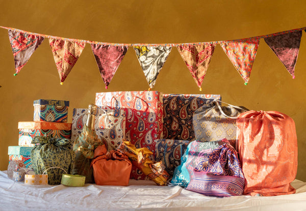 Colourful fabric bags and wrapped gifts on a table with a yellow wall and decorative banner in the background.