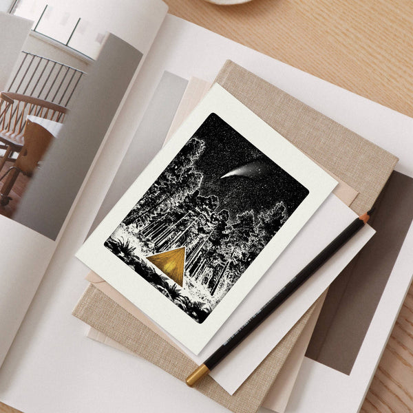 Black and white greeting card with a gold tent in a forest under a starry sky, on a desk with a pen.