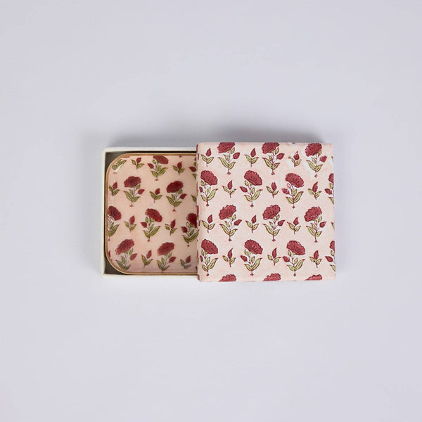A square decorative box with a floral print in muted red and olive green on a pale pink background, shown open with a matching patterned set of coasters inside against a light grey studio background.