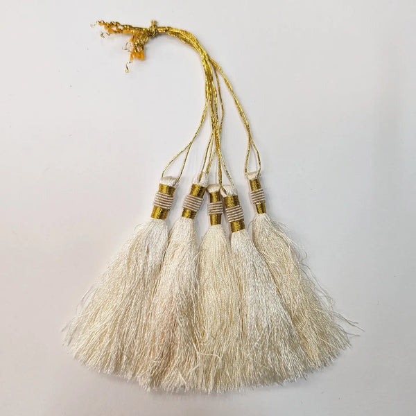 Five cream-coloured silk tassels with gold metallic bands and gold hanging threads, arranged against a plain off-white background.