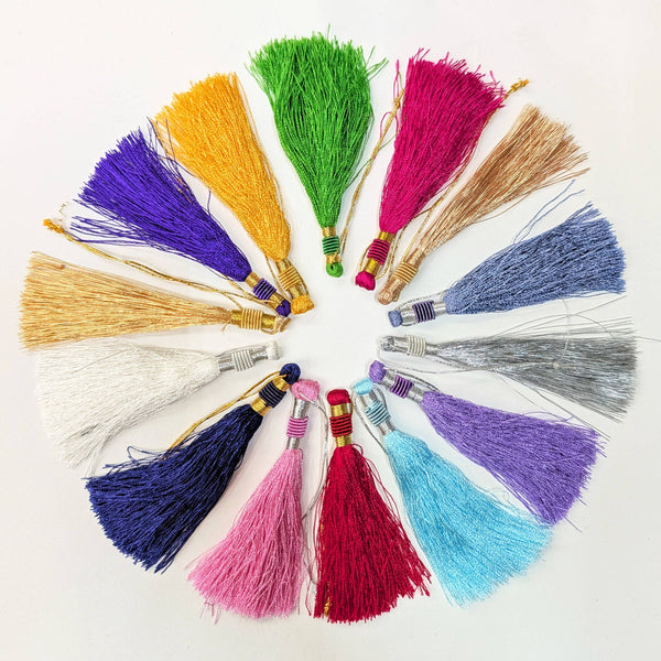 A set of colorful decorative tassels arranged in a circular pattern on a plain white background. The tassels come in multiple vibrant colors including green, pink, purple, yellow, blue, red, and white, each with a wrapped metallic cap.