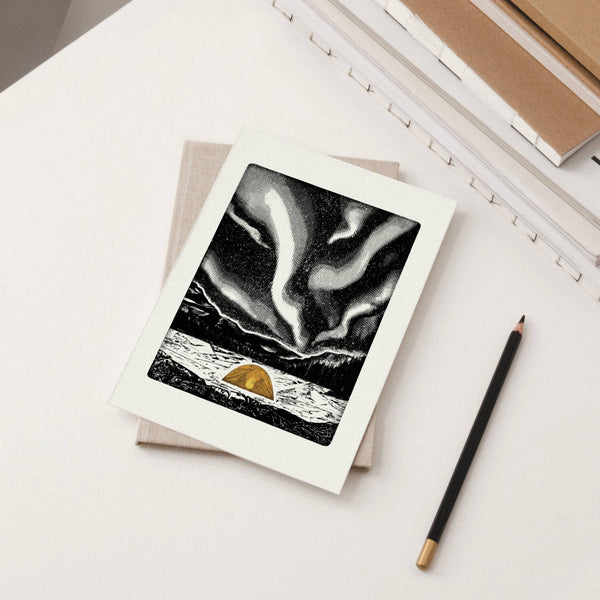 A white-bordered greeting card featuring a black-and-white northern lights scene with a small orange tent, styled on a white desk next to a pencil and stacked notebooks.