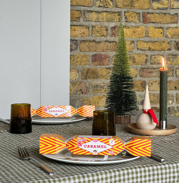 Two Christmas table settings with orange and yellow striped Tunnock's crackers, amber glass tumblers, a green candle, and a small artificial tree against a brick wall background.