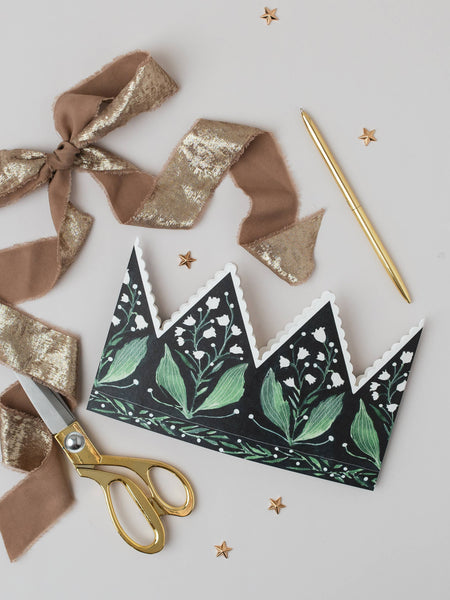 Decorative paper crown with green leaf design, gold scissors, and ribbons on a light gray background