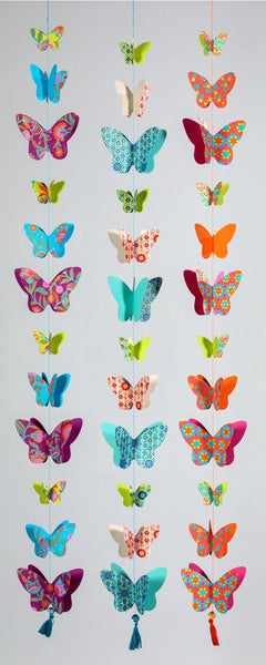 A vertical arrangement of colourful hanging butterfly decorations in many patterns and hues, suspended against a plain light grey background.