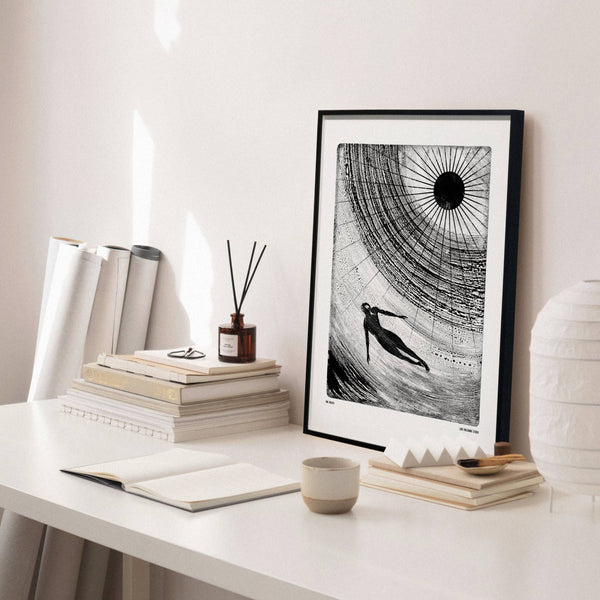 Framed black and white artwork on a desk with books and a lamp.