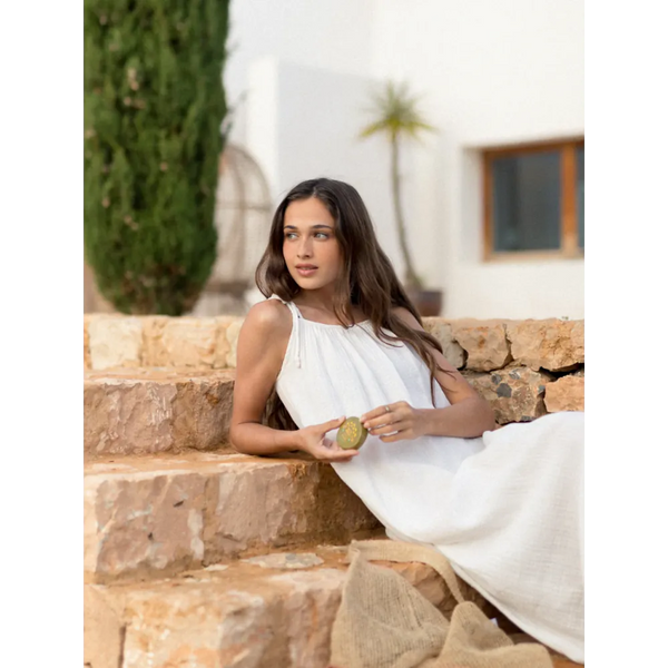 Woman in a white dress sitting on stone steps outdoors holding a bottle of Sol de Ibiza Face & Body Magic Sun Oil 