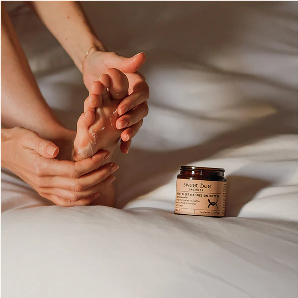 Person applying cream to their foot with a jar of 'sweet bee' magnesium butter on a bed.
